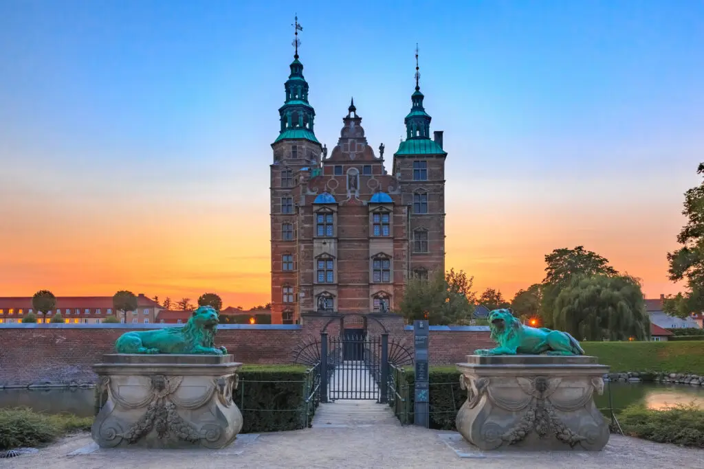 Sunset view of Rosenborg Castle in Copenhagen, Denmark, with bronze lion statues and an ornate gate leading to the historical building.