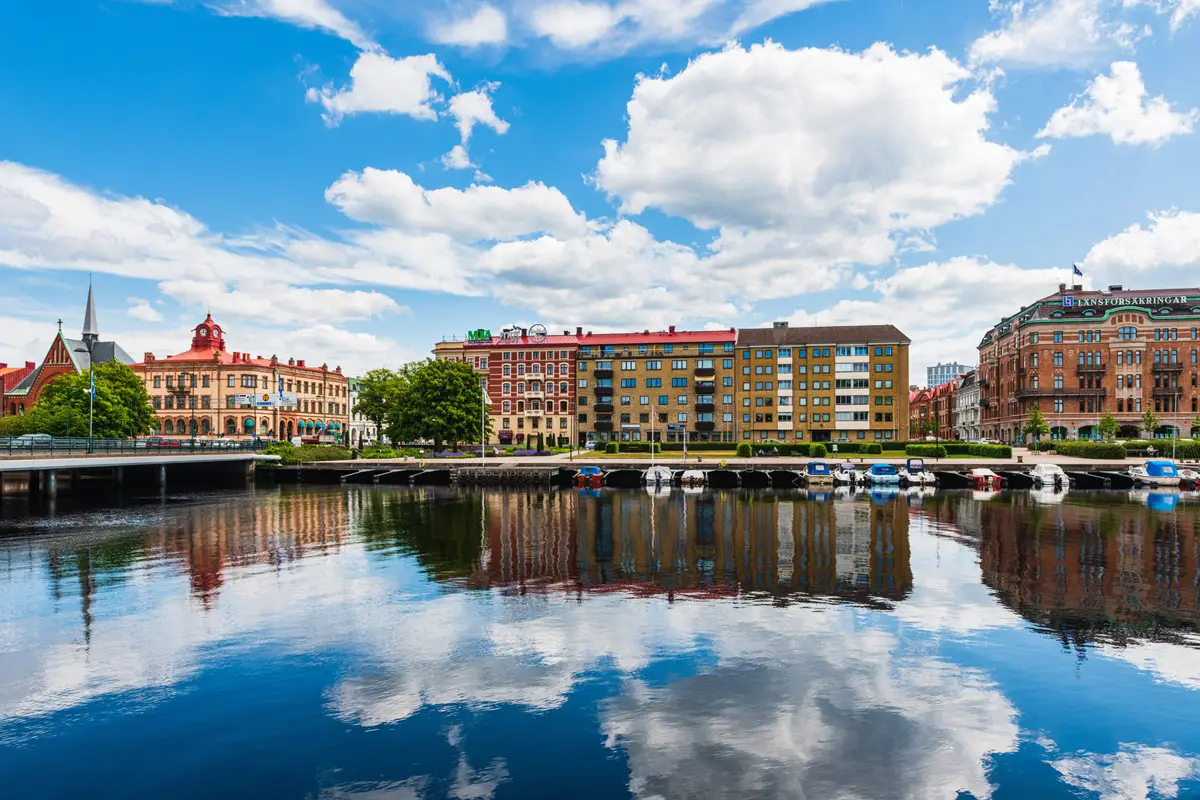 Colorful buildings lining the riverfront in Halmstad, Sweden, reflecting beautifully on the calm waters under a sunny blue sky with fluffy clouds.