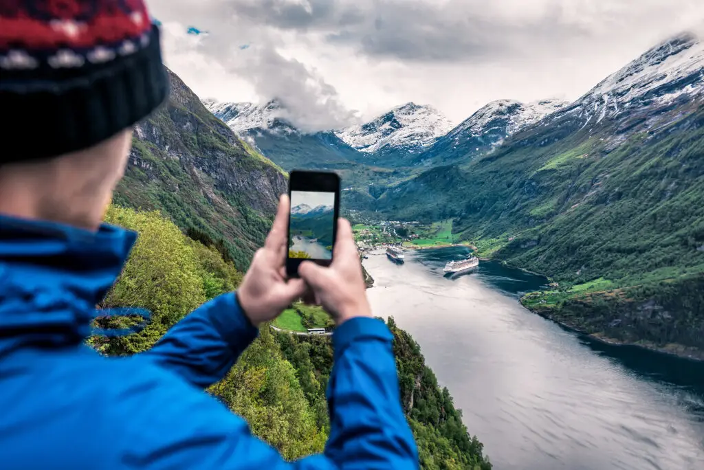 Tourist captures breathtaking view of Geiranger Fjord in Norway using smartphone, showcasing snow-capped mountains and serene waters with cruise ships.