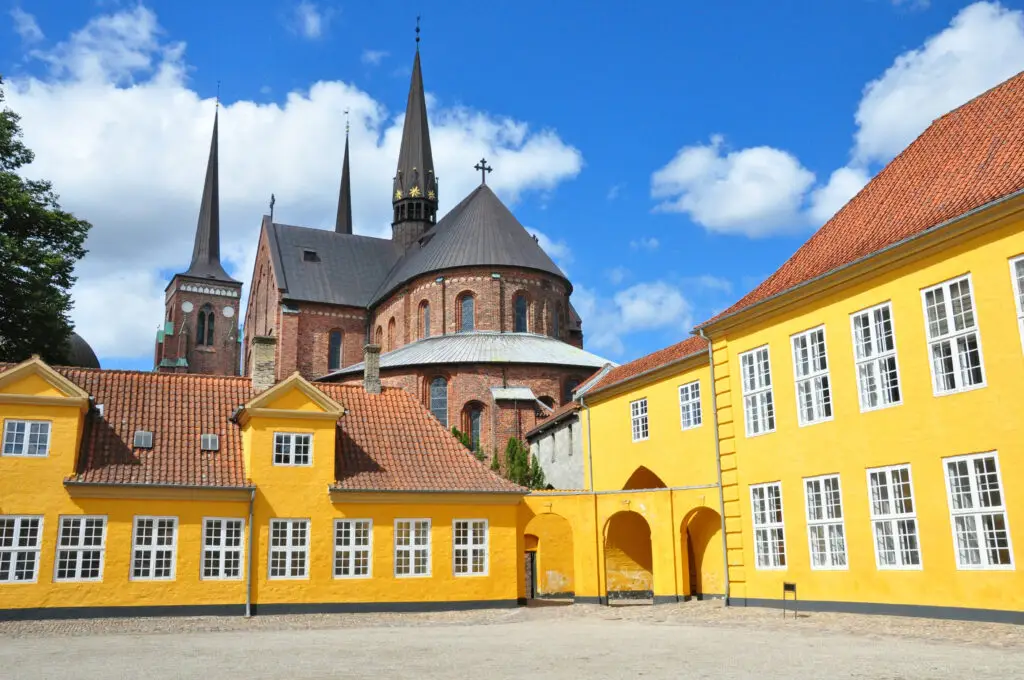 Roskilde Cathedral with its iconic twin spires behind the vibrant yellow buildings, a UNESCO World Heritage Site in Denmark, under a blue sky with fluffy clouds.