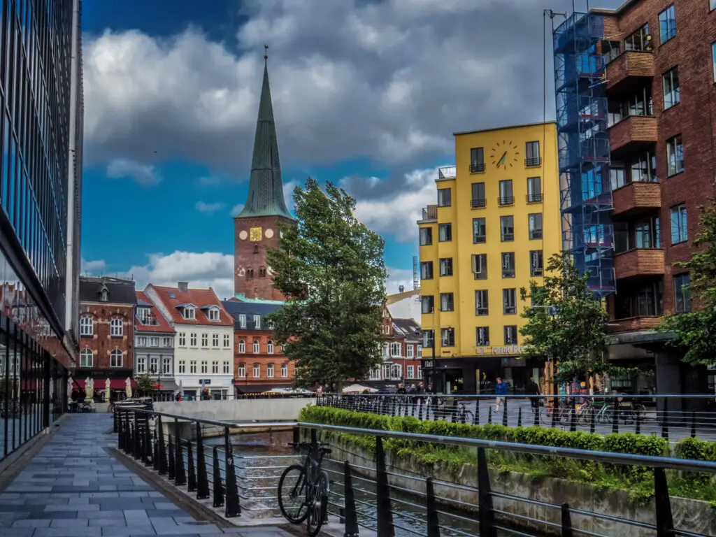 Colorful urban scene in Aarhus, Denmark showcasing a mix of traditional and modern architecture along a city water canal, highlighted by a prominent church spire and vibrant yellow building.
