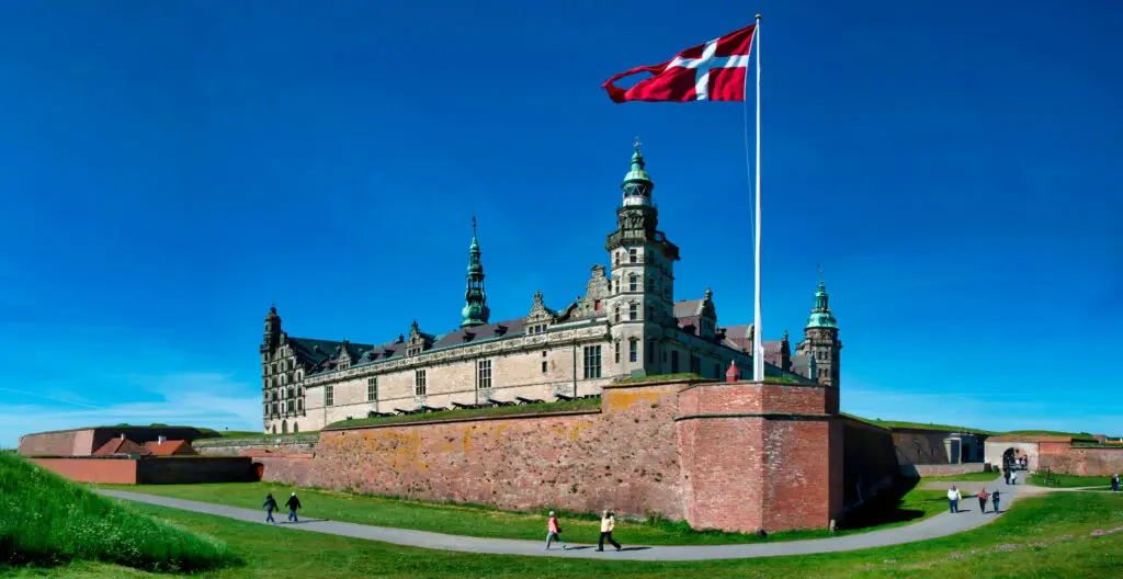 Kronborg Castle in Helsingør, Denmark under a clear blue sky, with the Danish flag waving and visitors exploring the historic site.