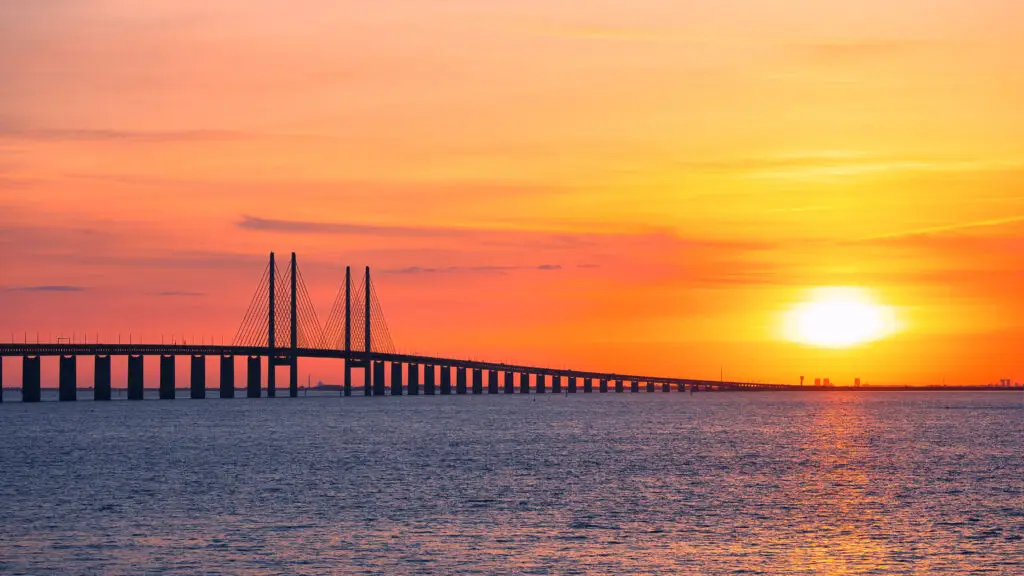 Sunset view of the Oresund Bridge connecting Malmo, Sweden, and Copenhagen, Denmark, showcasing the bridge's striking architecture against a vibrant orange sky.