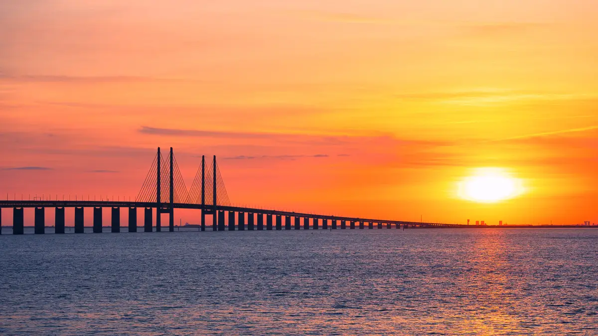 Sunset view of the Oresund Bridge connecting Malmo, Sweden, and Copenhagen, Denmark, showcasing the bridge's striking architecture against a vibrant orange sky.