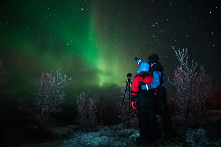 Two people observing the Aurora Borealis in Lapland, Sweden, capturing the vibrant northern lights above a snowy landscape.