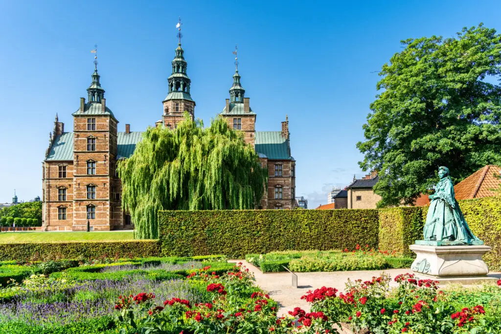 Statue of Queen Caroline Amalie overlooking lush gardens with vibrant flowers in the foreground and the historic Rosenborg Castle under a clear blue sky in Copenhagen, Denmark.