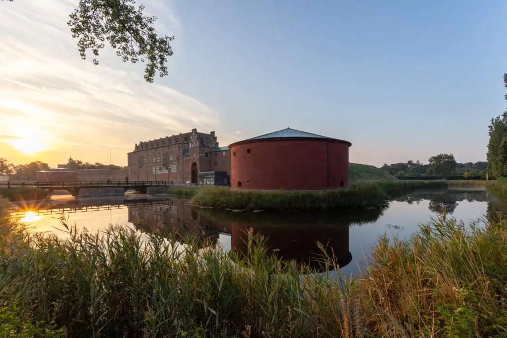 Sunrise view of the historic Castle of Malmö in Sweden, reflecting on the serene water surrounded by lush greenery.