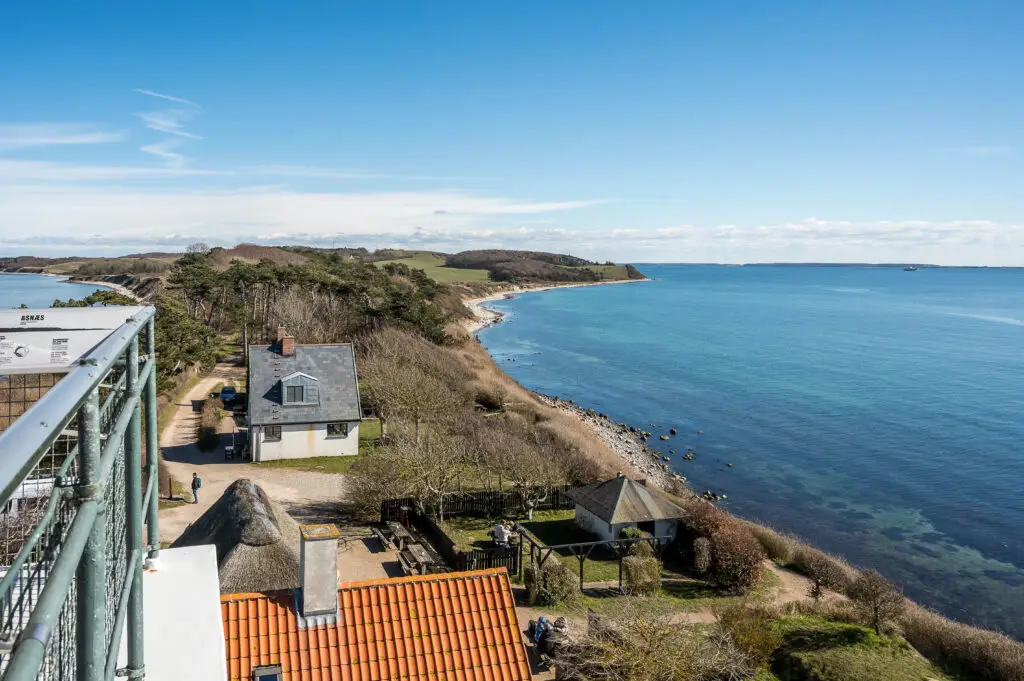 Scenic view from Røsnæs Lighthouse in Kalundborg, Denmark, showcasing the vast blue sea, coastal terrain, and a family relaxing in their backyard.