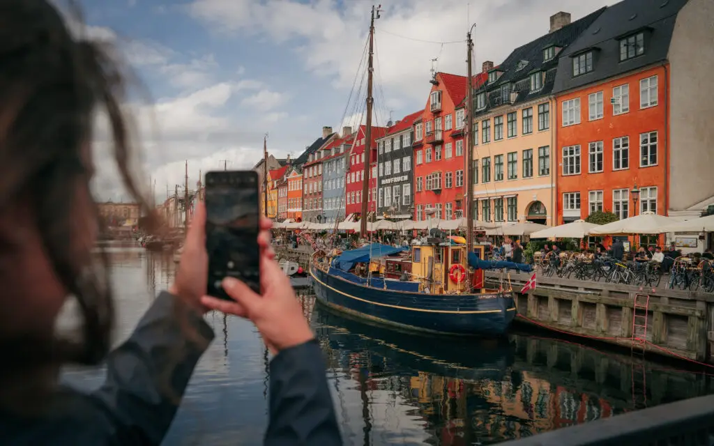 Woman using smartphone to capture scenic view of colorful buildings and boats at Nyhavn port, Copenhagen, enhancing the appeal for tourists and photographers.