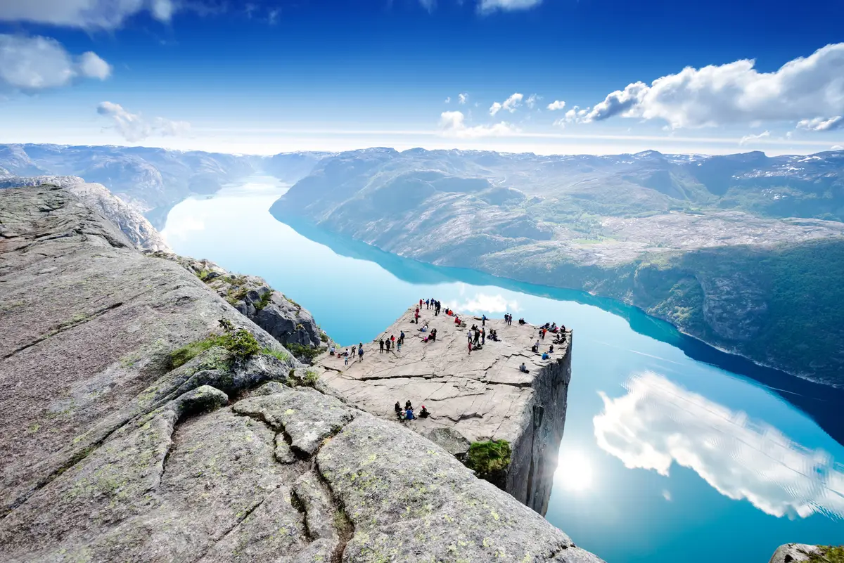 Tourists enjoying the breathtaking view from Preikestolen (Pulpit Rock) overlooking Lysefjord under a clear blue sky, a popular destination in Norway for hiking and nature tours.