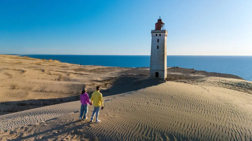Couple walking toward iconic white lighthouse on sandy shores of Denmark under clear blue sky