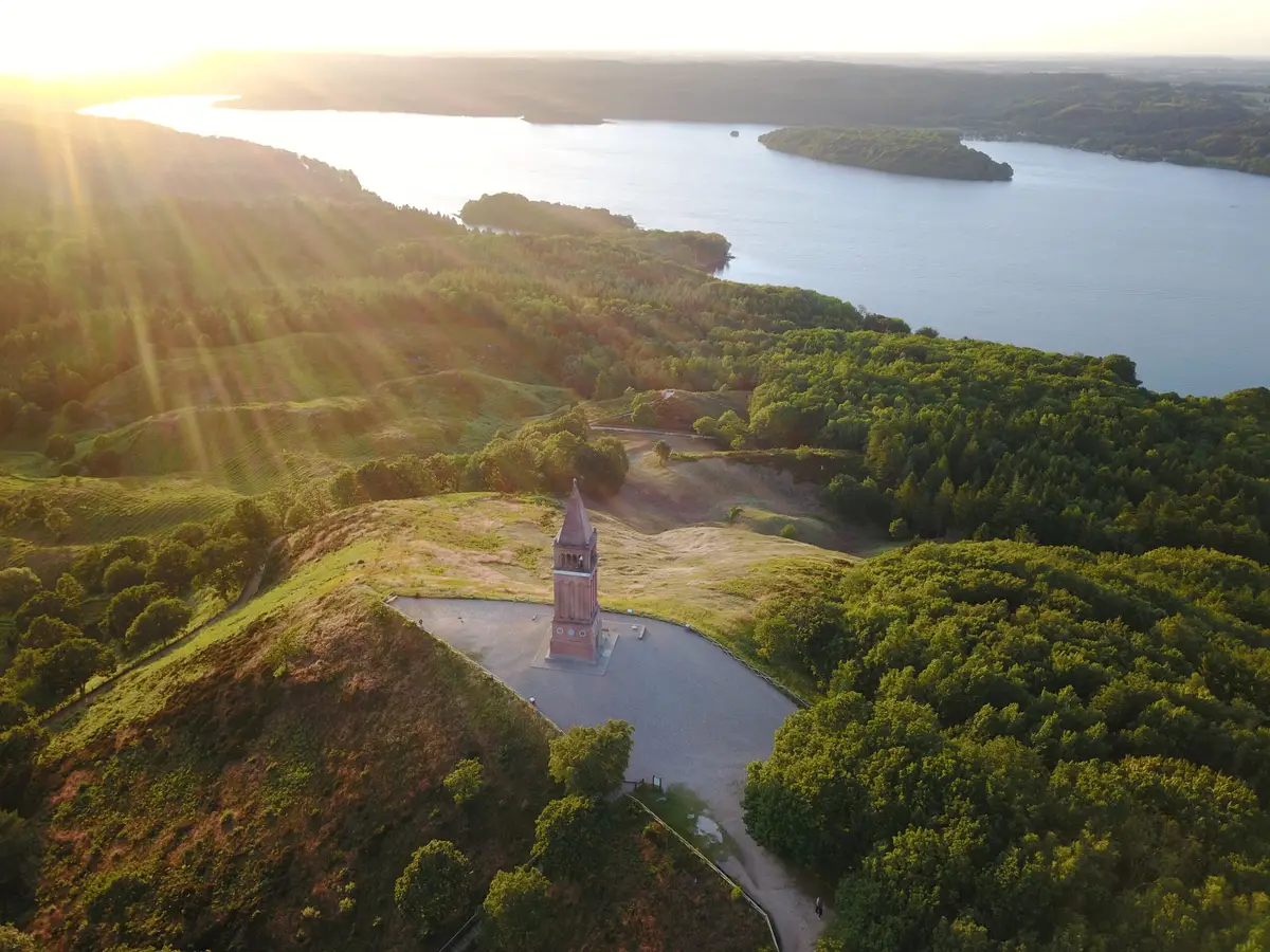 Aerial view of Himmelbjerget with its iconic tower surrounded by lush forests and the serene Julso Lake during sunset, a popular tourist attraction in Denmark.