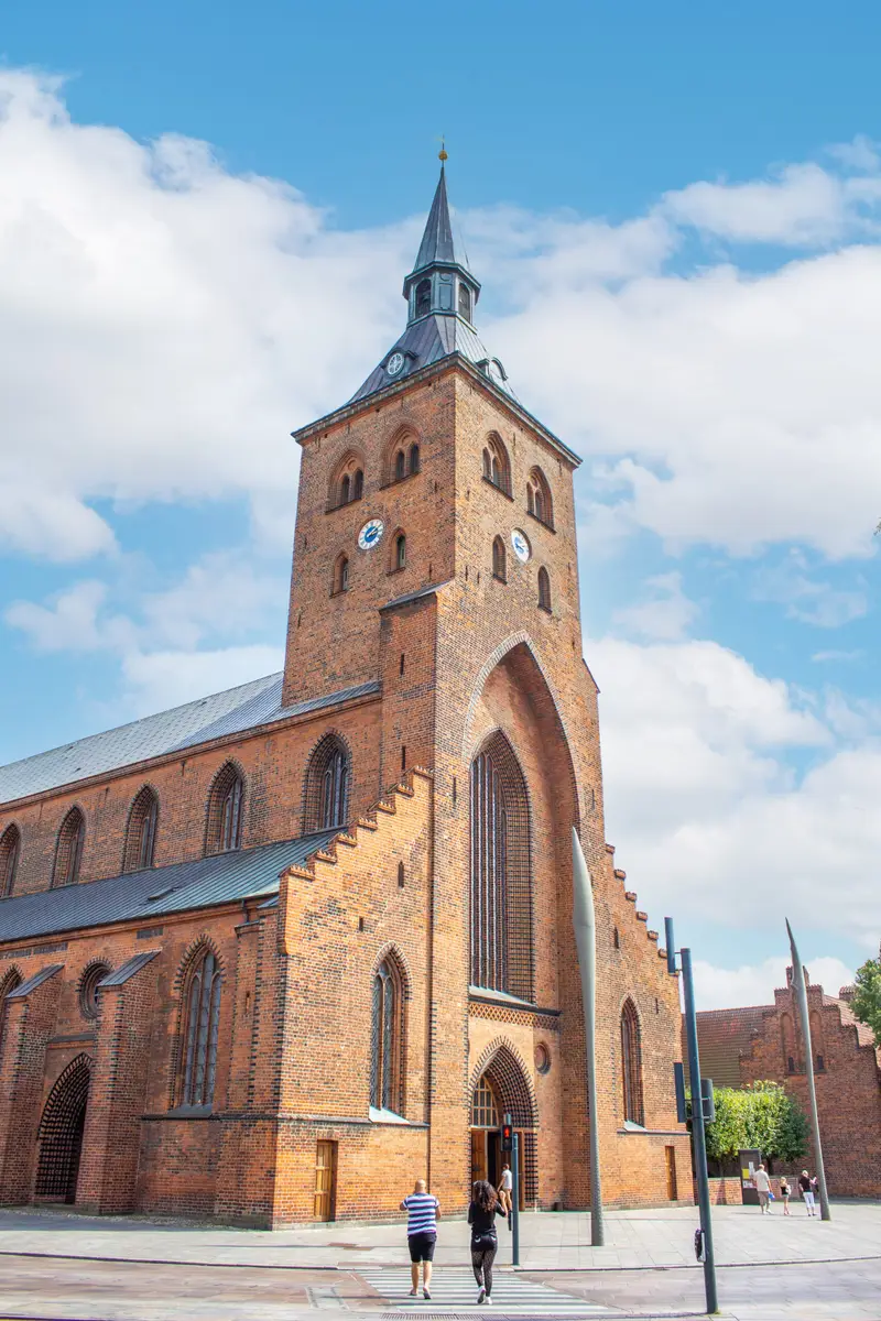 Tourists walking towards the historic Sankt Knuds Kirke in Odense, Denmark, showcasing its distinctive Gothic architecture under a blue sky.
