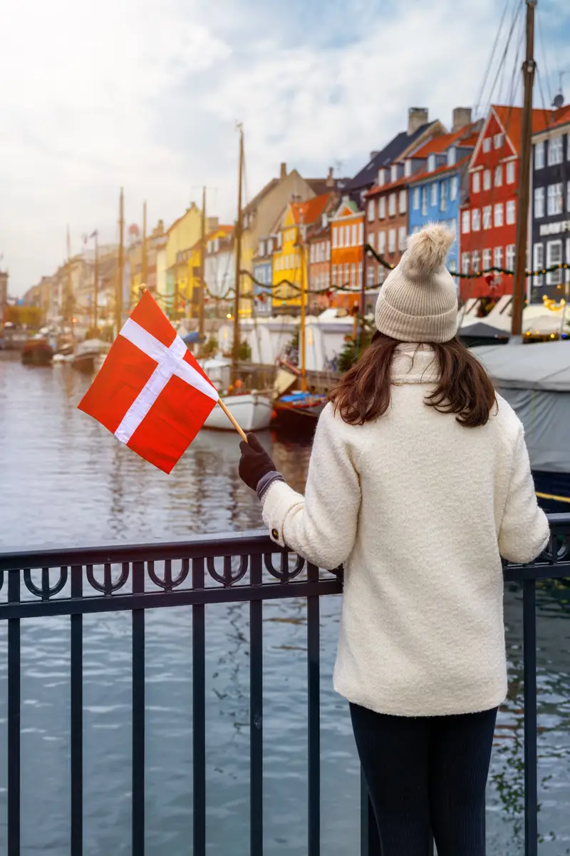 Tourist woman holding a Danish flag while admiring the colorful waterfront buildings of Nyhavn in Copenhagen, Denmark during winter