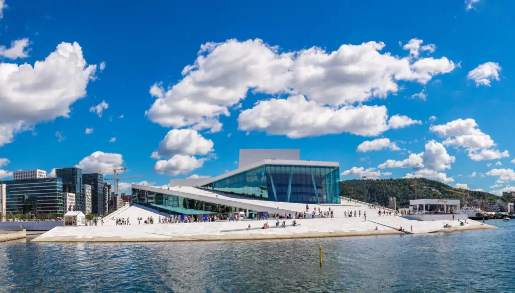 View of the Oslo Opera House in Norway, showcasing its modern architecture and bustling waterfront with visitors and clear blue skies.