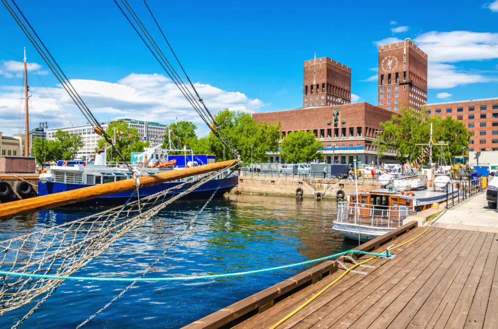 Oslo City Hall viewed from the harbour on a sunny day, featuring historic boats and clear blue waters, Norway.